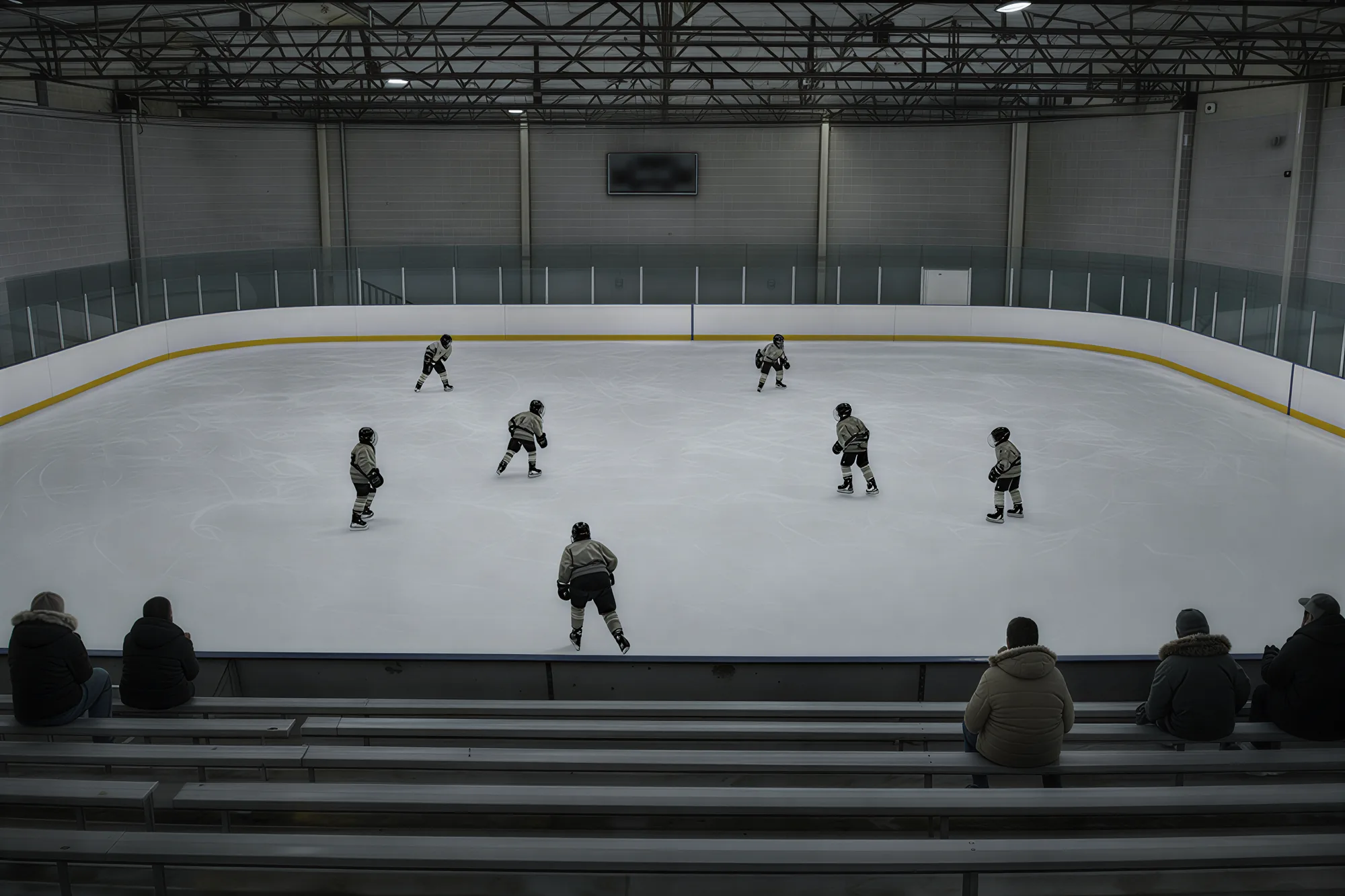 A small indoor rink from the stands: concrete block walls, steel roof trusses, a dark scoreboard, youth players on the ice and a few parents on the benches below.