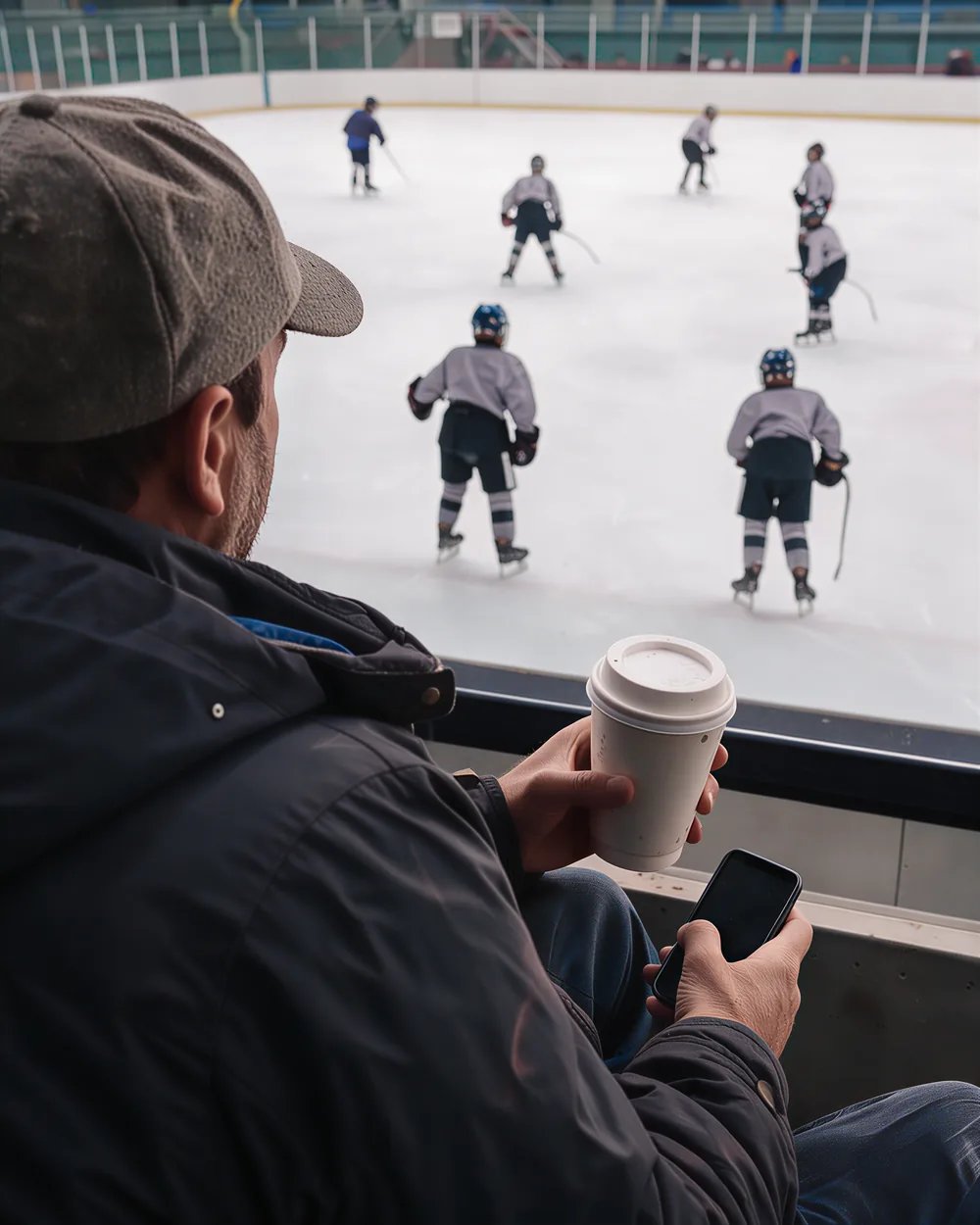 A father in a cap seen from behind in the stands, a paper coffee cup in one hand and a phone low in the other, youth hockey players on the ice beyond the glass.