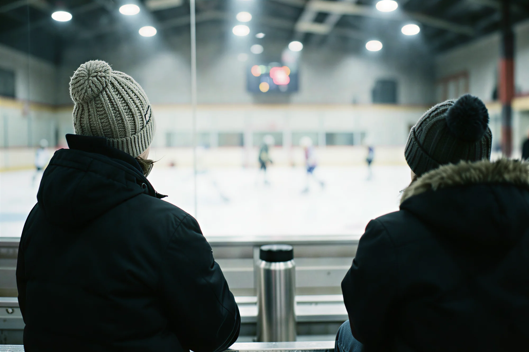 Two parents in knit hats watch a youth hockey game from the cold bleachers, a thermos between them, the game soft behind the glass.