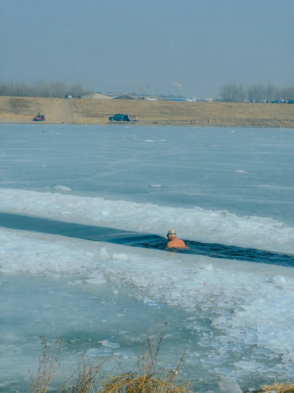 A swimmer in a channel cut through the ice of a frozen lake, seen from the shore.